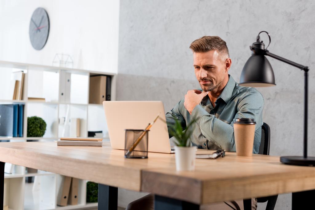 pensativo hombre de negocios sentado en el escritorio y mirando a la computadora portátil en la oficina moderna
 - Foto, Imagen
