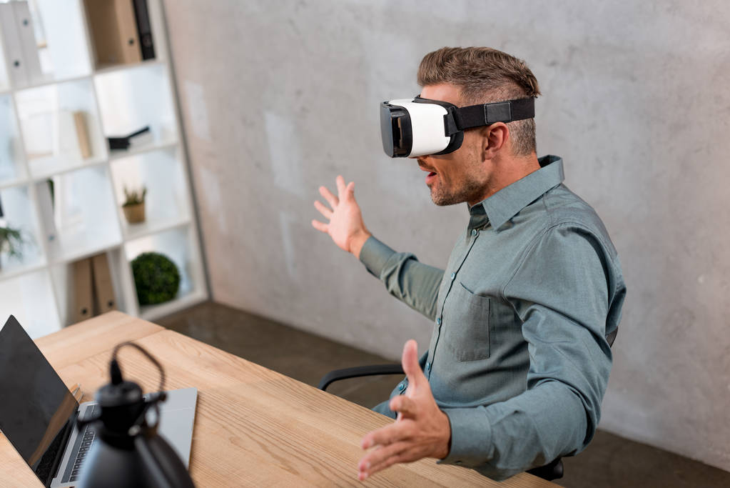 オフィスでラップトップのそばに座ってながら仮想現実のヘッドセットを着てびっくり男  - 写真・画像
