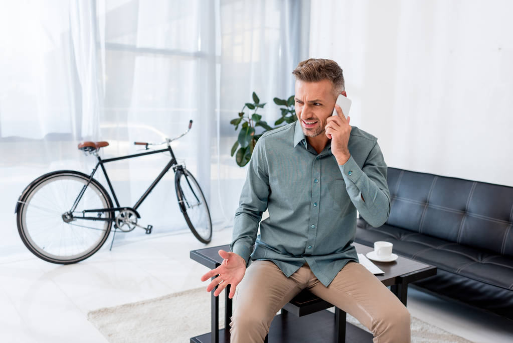 homem de negócios emocional falando no smartphone enquanto sentado na mesa de café no escritório
  - Foto, Imagem