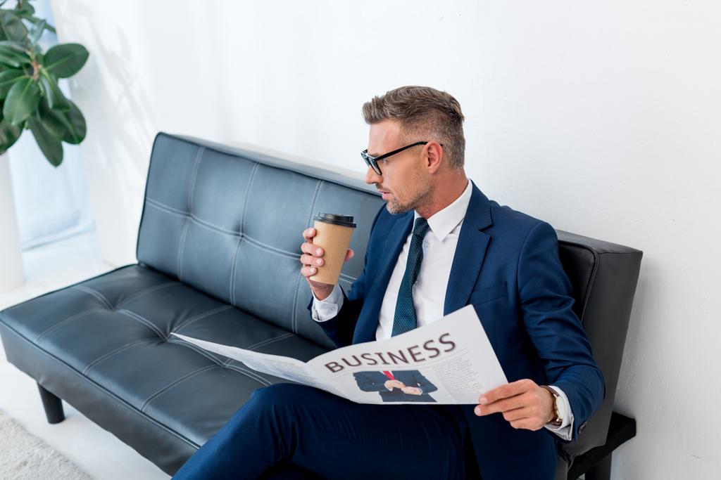 hombre de negocios en traje sosteniendo periódico de negocios y taza de papel mientras está sentado en el sofá
  - Foto, Imagen