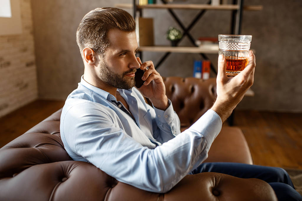 Молодой привлекательный бизнесмен сидит на диване и смотрит на стакан виски в руке в собственном кабинете. Парень разговаривает по телефону. Серьезный и сосредоточенный. Сексуальный молодой человек
. - Фото, изображение