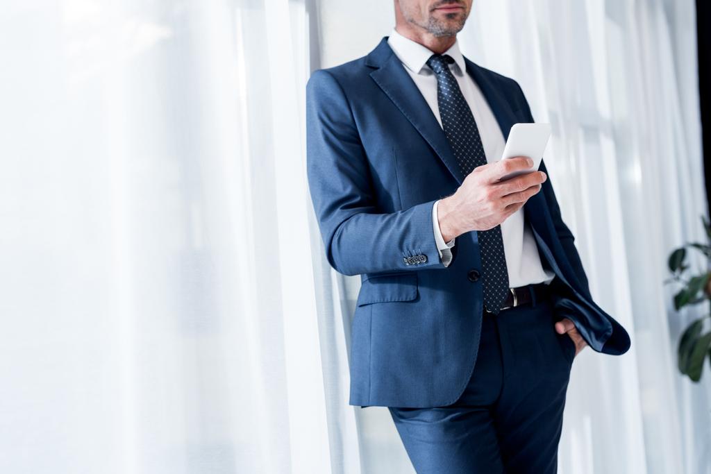 vista recortada del hombre de negocios en traje de pie con la mano en el bolsillo mientras se utiliza el teléfono inteligente
 - Foto, Imagen
