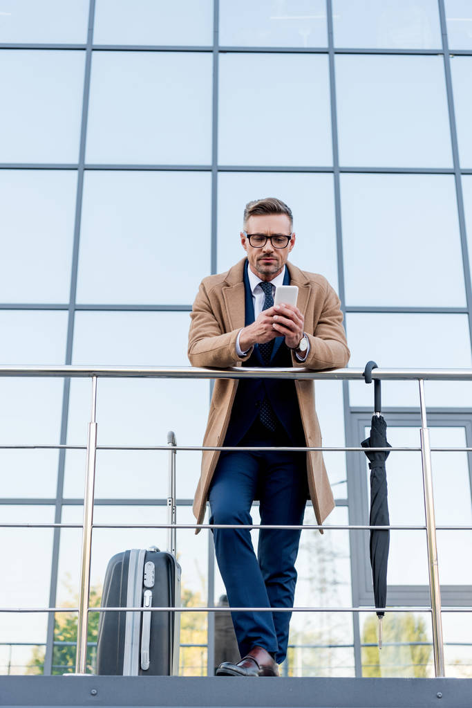 guapo hombre de negocios con abrigo beige usando un teléfono inteligente mientras está de pie cerca de la maleta y el paraguas
  - Foto, Imagen