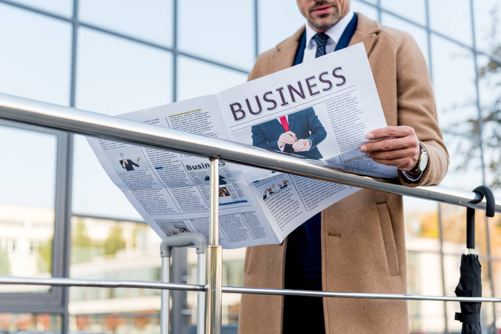Ausgeschnittene Ansicht eines Geschäftsmannes, der im beigen Mantel steht und Wirtschaftszeitung liest  - Foto, Bild