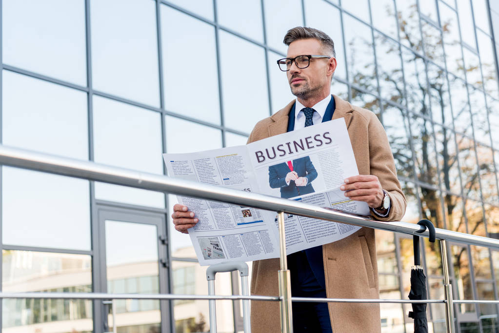 komea liikemies seisoo beige takki ja tilalla liike sanomalehti
  - Valokuva, kuva