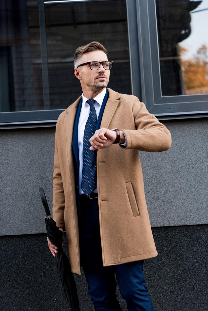 handsome businessman standing with watch on hand and holding umbrella  - Photo, Image