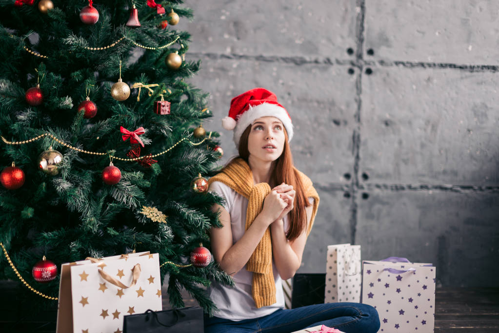 испуганная серьезная девушка сидит под новогодней елкой
 - Фото, изображение