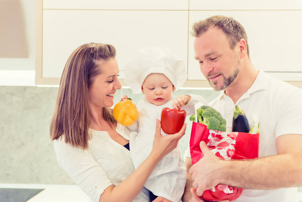 Happy parents standing in the kitchen and prepares for cooking with their baby girl chef. Father bring vegetables in paper bag. - Photo, Image