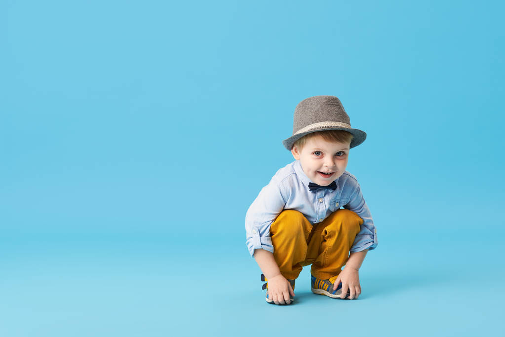 Portret van gelukkig vreugdevolle jongetje geïsoleerd op blauwe achtergrond. Peuter kind in hoed en modieuze pak glimlachen en hebben een leuke  - Foto, afbeelding