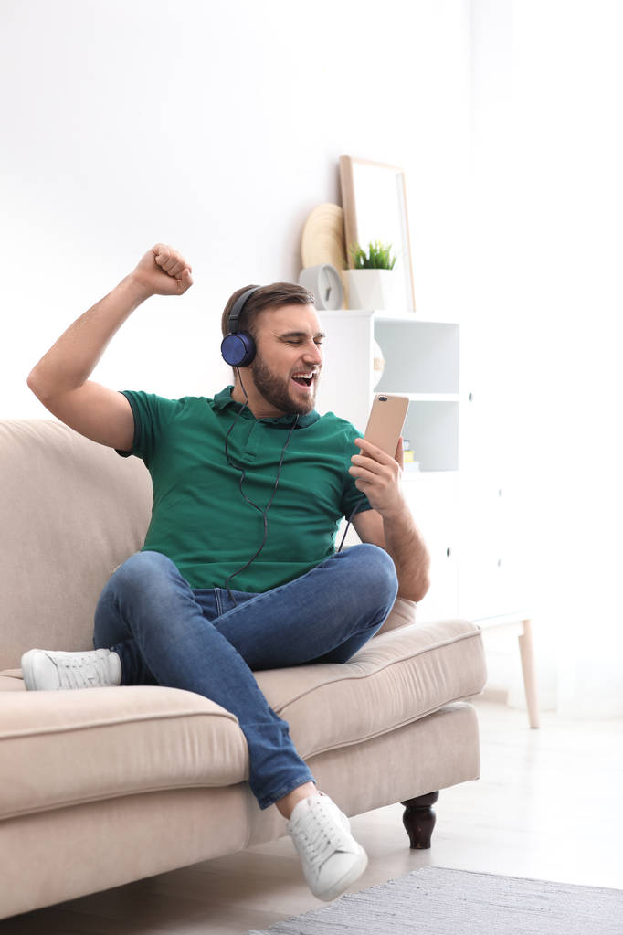 Молодой человек в наушниках с мобильным устройством наслаждается музыкой на диване дома
 - Фото, изображение