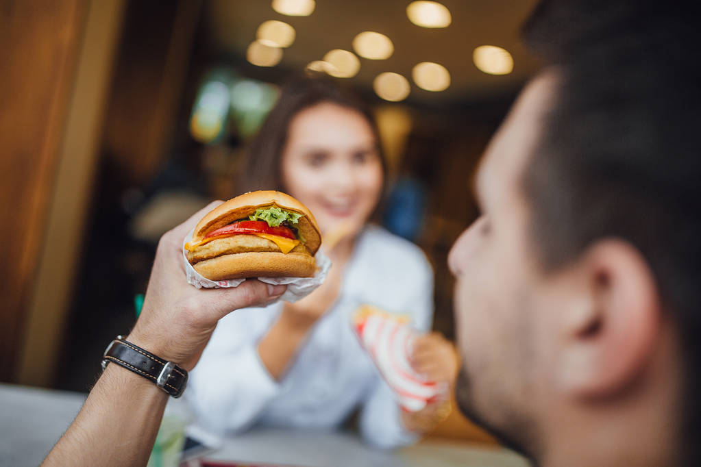 Joven hombre caucásico sabroso comer hamburguesa con queso en el fondo de la mujer, enfoque selectivo
 - Foto, Imagen