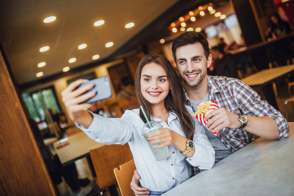 pareja joven haciendo selfie en el teléfono móvil con mojito y hamburguesa, sonriendo en la cafetería de comida rápida, enfoque selectivo
 - Foto, imagen