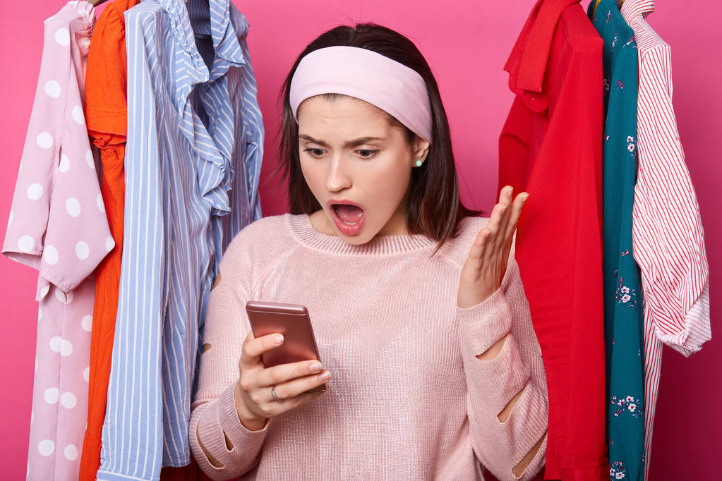 Shocked woman looks at smartphone display. Interesting information on screen. Dark haired lady raises palm and looks surprisingly at her gadget. Lots clothes behind beautiful female in shopping mall. - Photo, Image