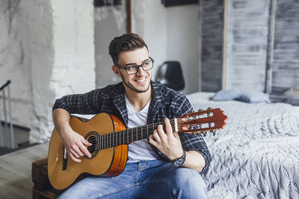 Молодий красивий чоловік в окулярах посміхається і позує з гітарою на ліжку
  - Фото, зображення