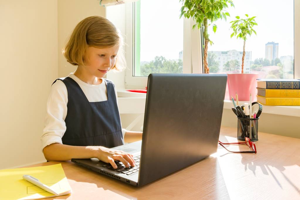 Little schoolgirl uses computer laptop sitting at a desk at home. School, education, knowledge and children - Photo, Image