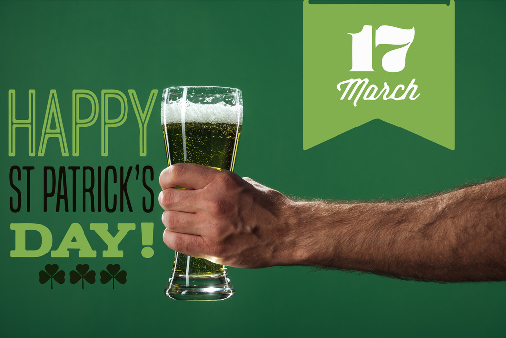 abgeschnittene Ansicht eines Mannes, der ein Glas Bier mit Schaum in der Nähe von happy st patricks day Schriftzug auf grünem Hintergrund hält - Foto, Bild