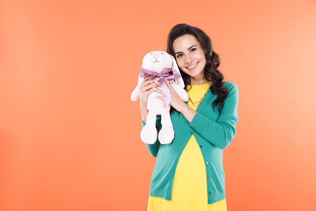 Feliz mujer embarazada rizada sosteniendo juguete conejo aislado en naranja
 - Foto, imagen