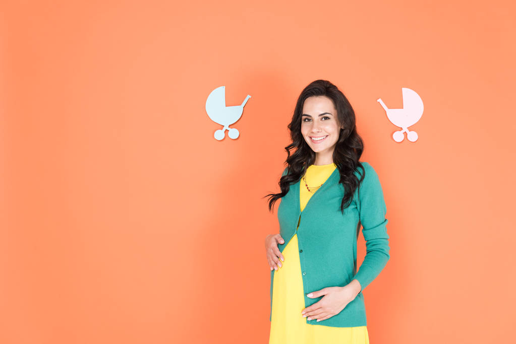 Excited brunette pregnant woman in green cardigan touching belly on orange background - Photo, Image