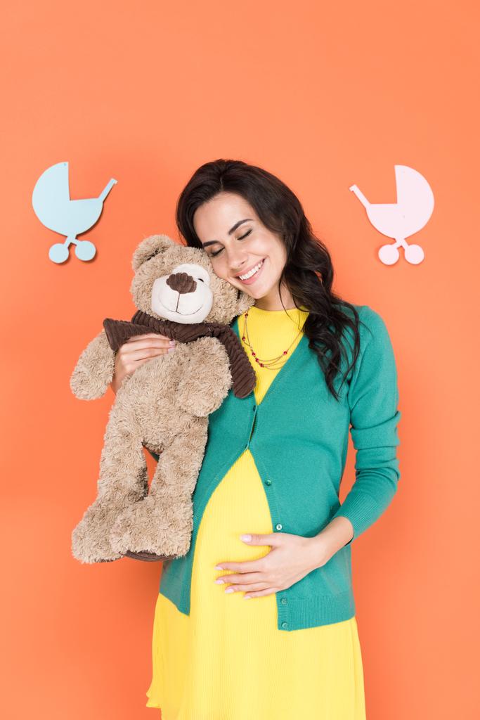 glücklich schwangere Frau mit Teddybär und anrührendem Bauch mit geschlossenen Augen auf orangefarbenem Hintergrund - Foto, Bild