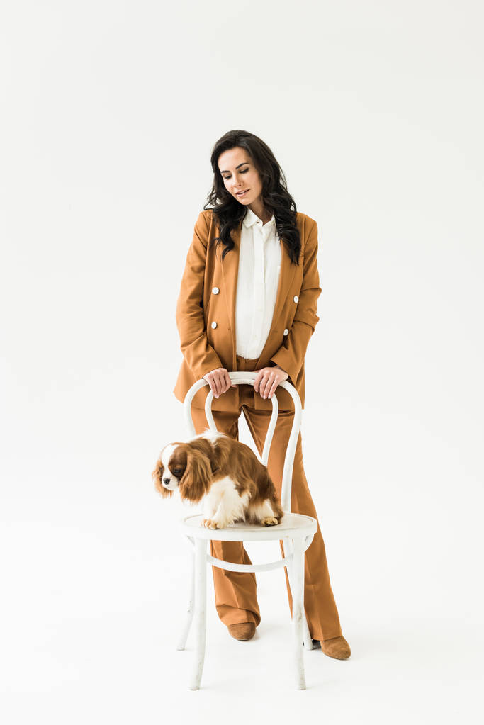 Stylish pregnant woman in suit looking at dog on chair on white background - Photo, Image