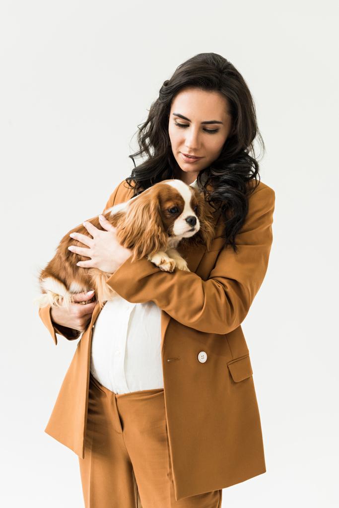 lockige schwangere Frau im braunen Anzug hält Hund isoliert auf weißem Grund - Foto, Bild