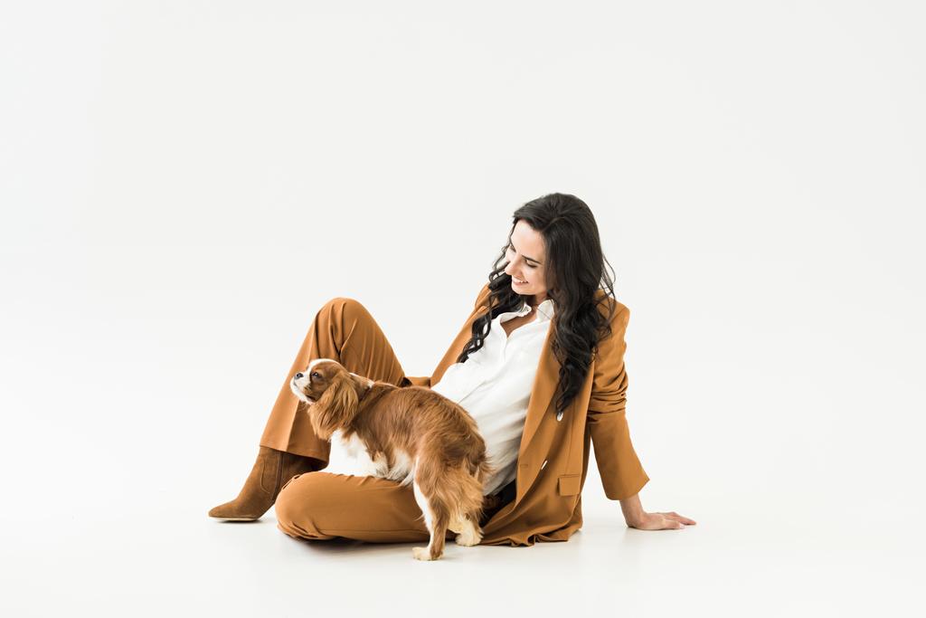 Gelukkig zwangere vrouw in pak zittend op de vloer met hond op witte achtergrond - Foto, afbeelding