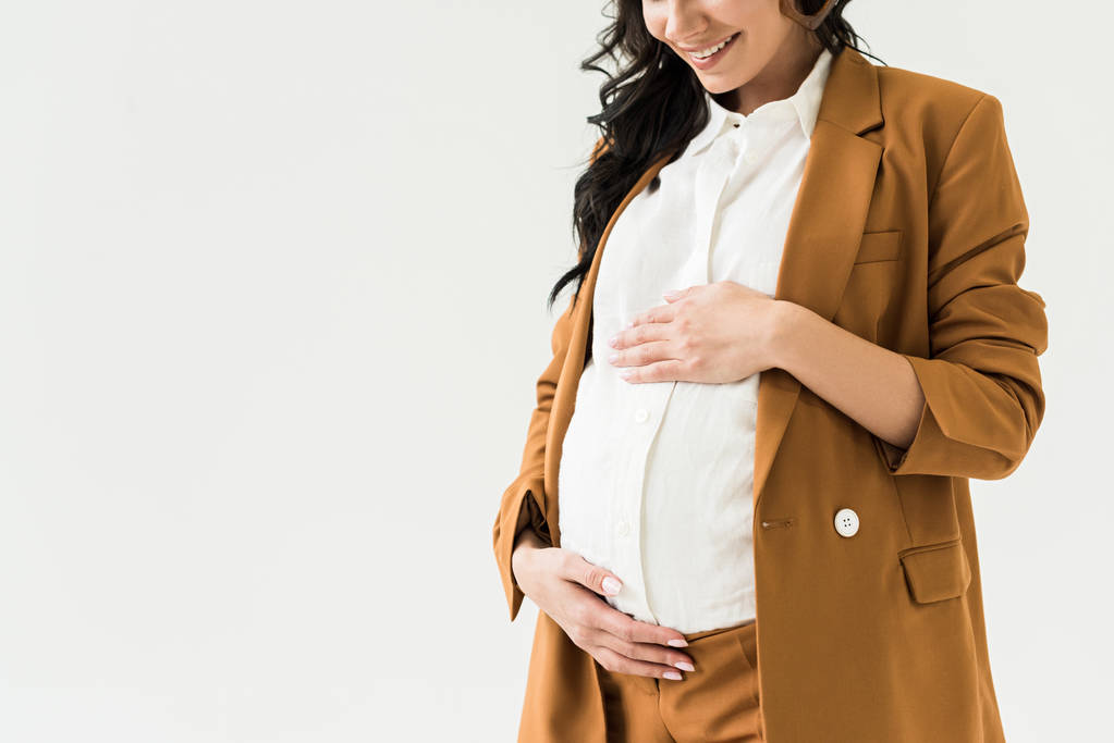 Vista recortada de la mujer embarazada riendo en traje marrón tocando vientre aislado en blanco
 - Foto, Imagen