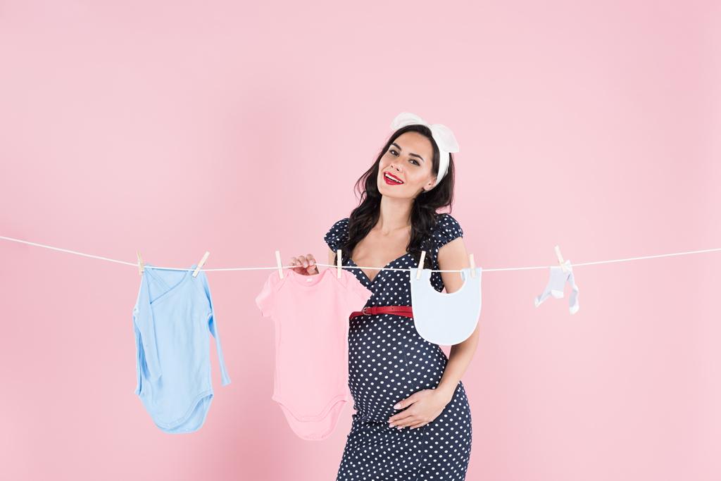 Terhes nő pöttyös ruha lógott ki a baba ruhákat a szárítókötél a rózsaszín háttér - Fotó, kép