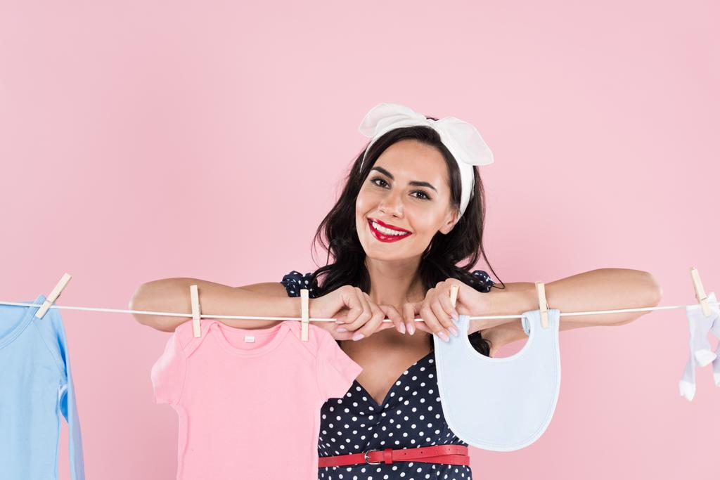 Mooie brunette jonge vrouw opknoping kleren van de baby op de waslijn geïsoleerd op roze - Foto, afbeelding