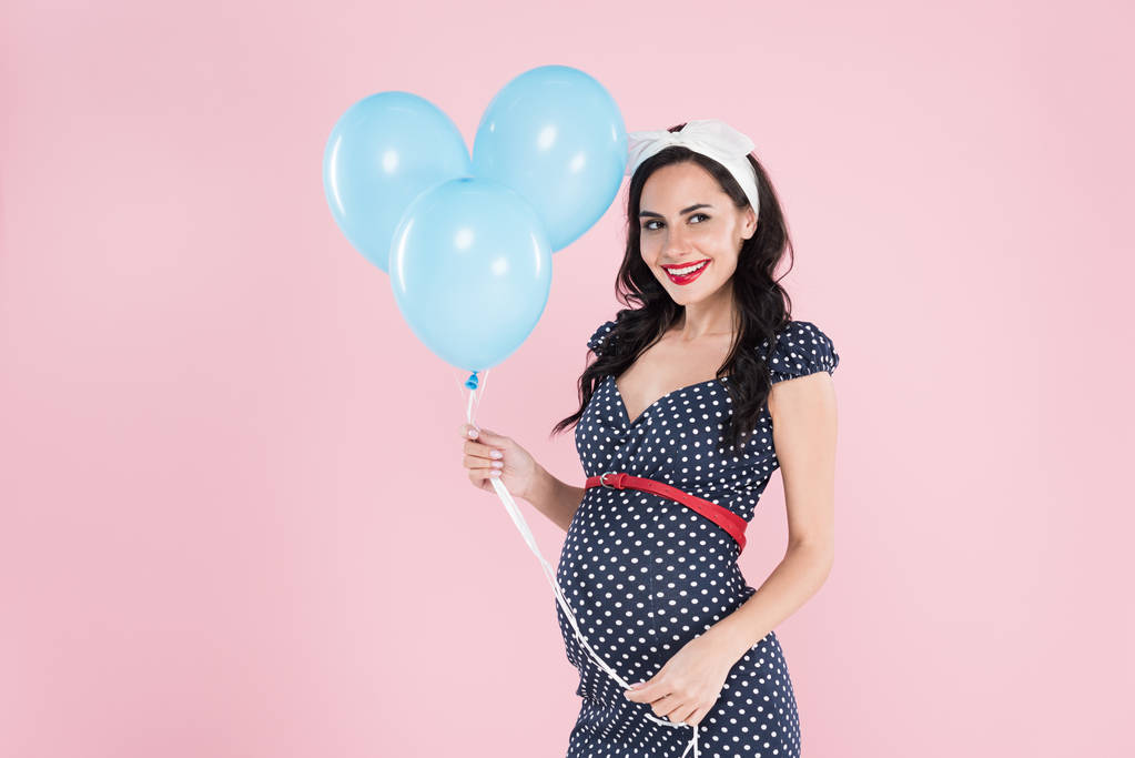 Щаслива вагітна жінка в точковій сукні Полки, що тримає блакитні повітряні кульки ізольовані на рожевому
 - Фото, зображення