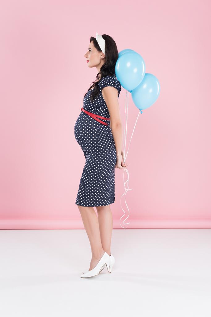 Side view of pregnant woman in dotted dress holding air balloons on pink background - Photo, Image