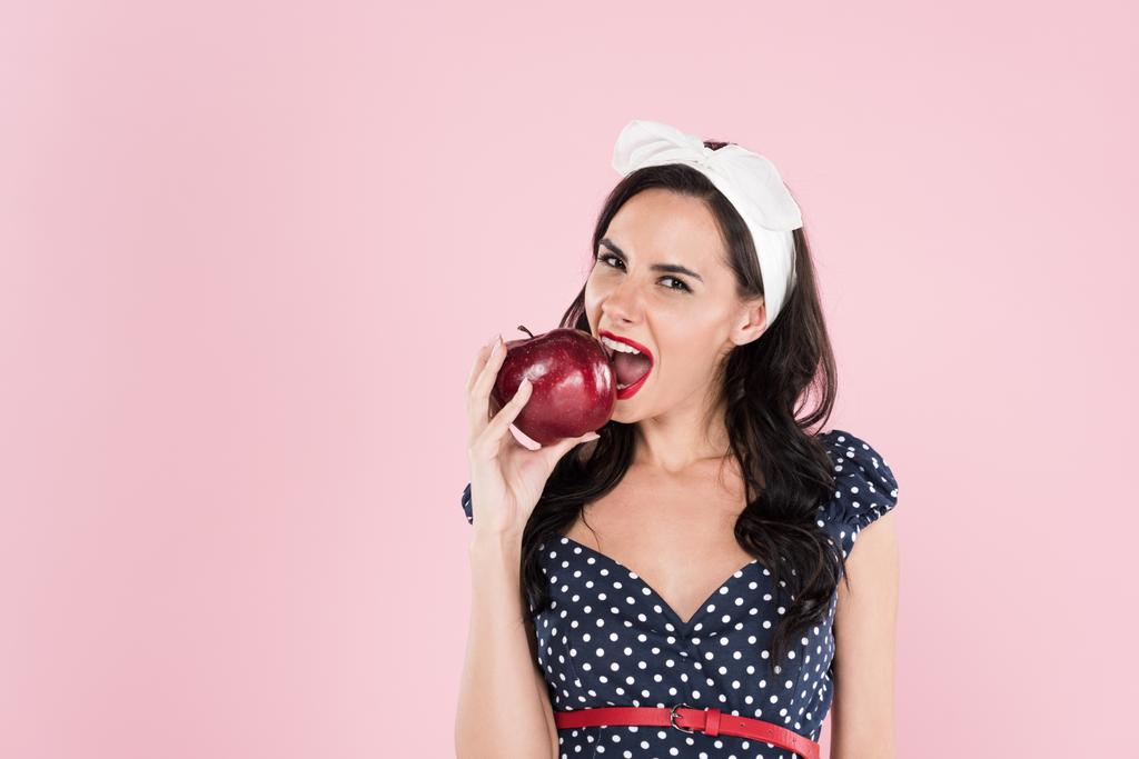 Linda menina morena em vestido de bolinhas comendo maçã vermelha isolada em rosa
 - Foto, Imagem