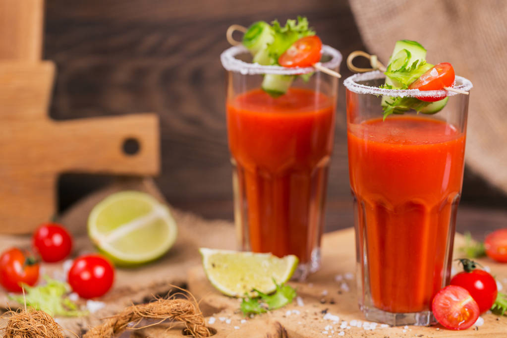 Дві склянки томатного соку прикрашені свіжими помідорами, огірком та листям на дерев'яному фоні
 - Фото, зображення