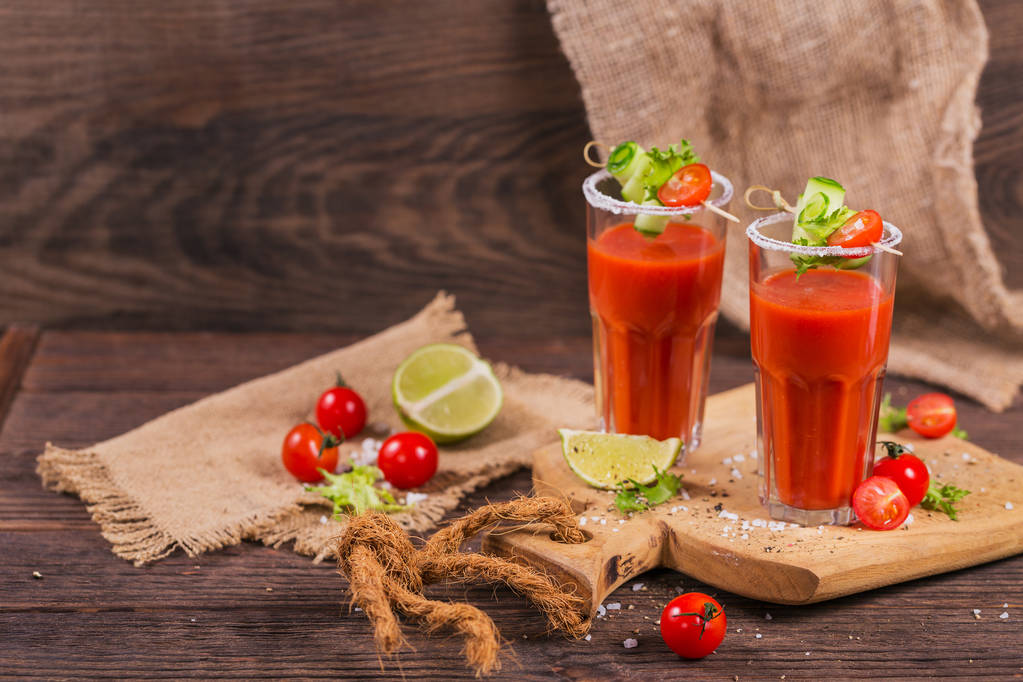 Два стакана томатного сока, украшенные свежими помидорами, огурцом и листьями на деревянном фоне
 - Фото, изображение