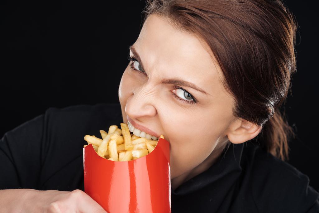 emocionální žena jíst chutné hranolky při pohledu na fotoaparát izolované na černém pozadí - Fotografie, Obrázek