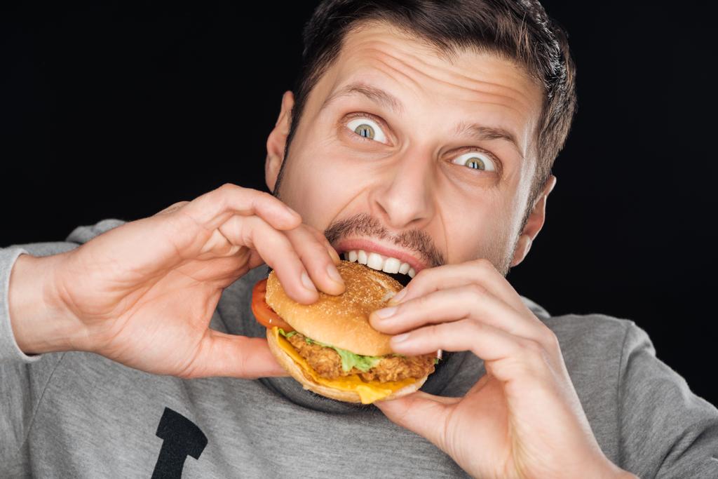 emocjonalne mężczyzna jedzenie smaczne kurczaka burger patrząc na kamery na czarnym tle - Zdjęcie, obraz