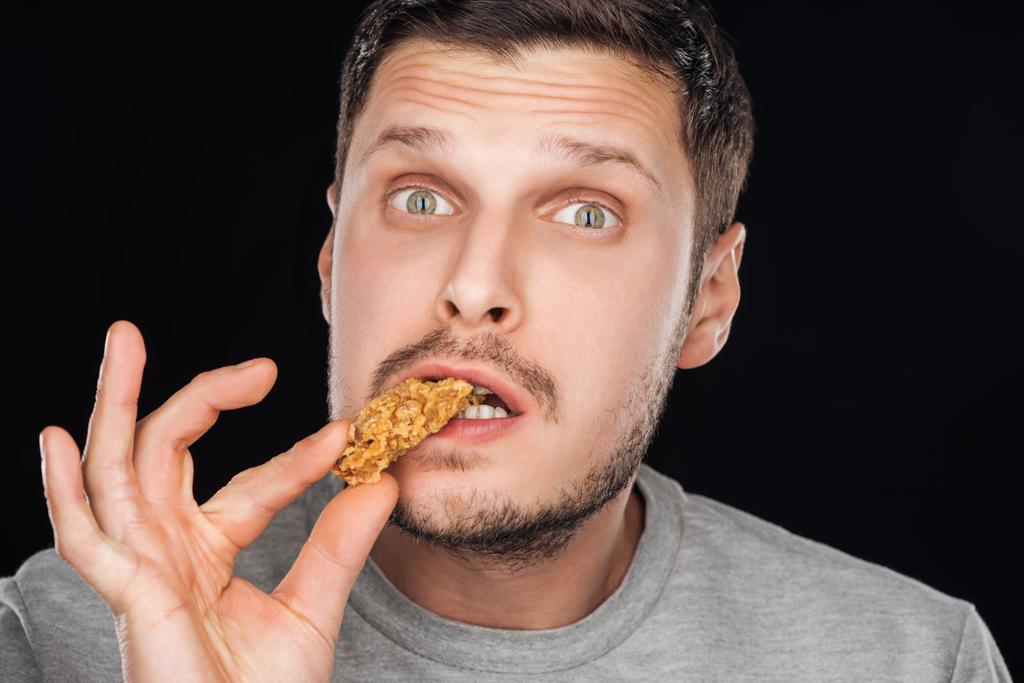 handsome man eating chicken nugget while looking at camera isolated on black - Photo, Image