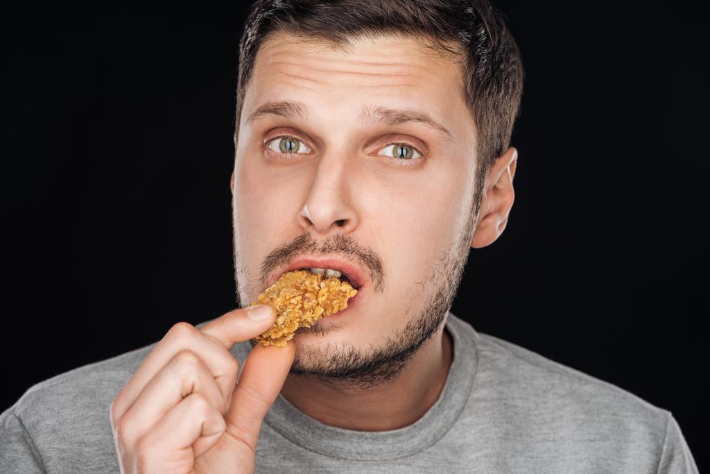 pohledný muž, jíst chutné kuřecí nugget při pohledu na fotoaparát izolované na černém pozadí - Fotografie, Obrázek