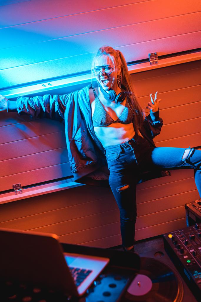 selektiver Fokus eines glücklichen blonden DJ-Mädchens mit Brille, das im Nachtclub steht und gestikuliert - Foto, Bild