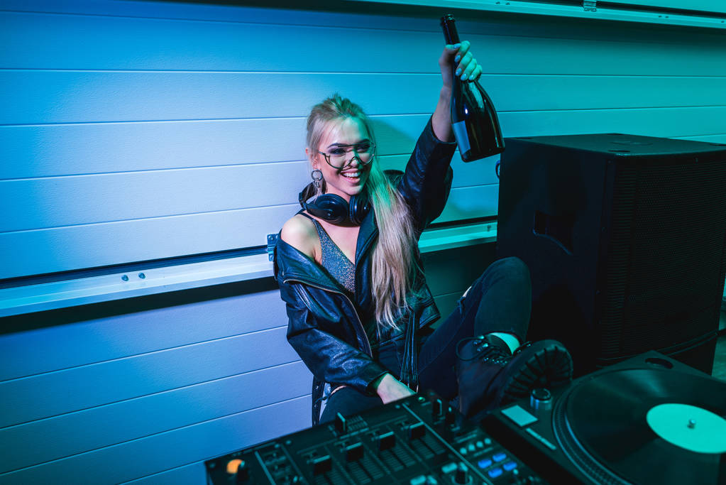 fröhliches DJ-Mädchen hält Flasche mit Wein in der Hand und sitzt im Nachtclub - Foto, Bild