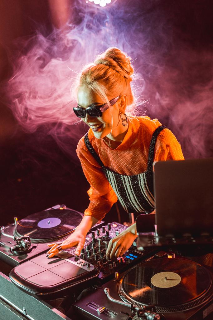 dj veselá dívka v sluneční brýle dotýká dj vybavení v nočním klubu s kouřem  - Fotografie, Obrázek