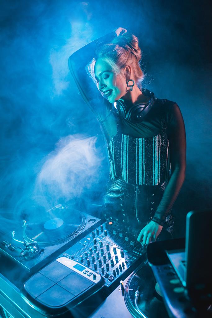 glückliches blondes DJ-Mädchen mit Kopfhörern, das in der Nähe von DJ-Geräten in einem Nachtclub mit Rauch steht  - Foto, Bild