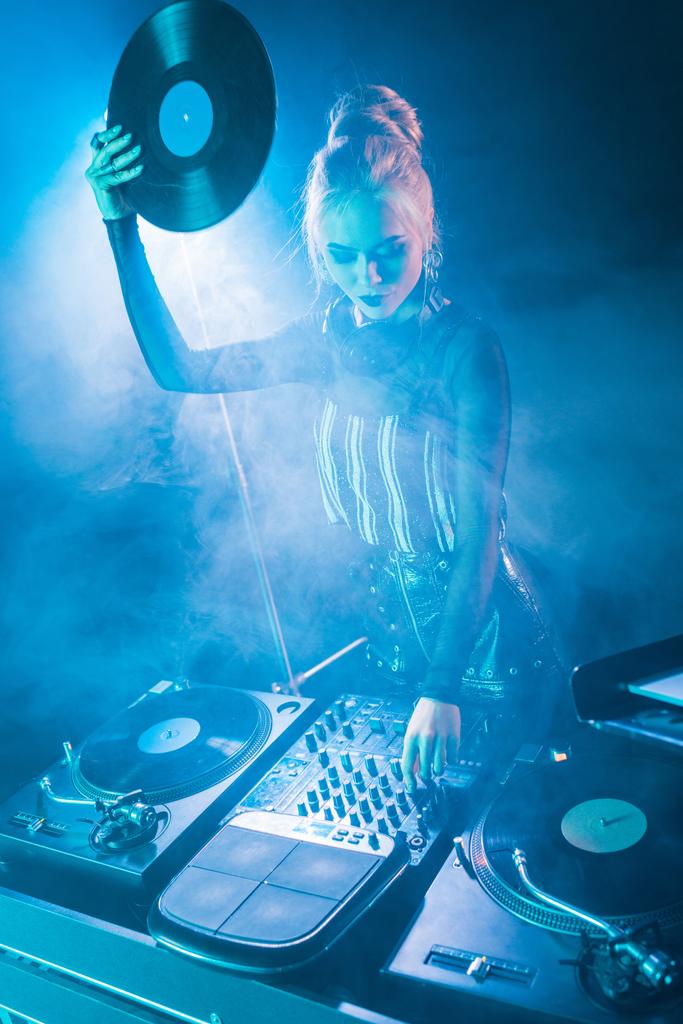 blonde dj meisje kijkend naar dj apparatuur en houden van retro vinyl record in nachtclub met rook  - Foto, afbeelding