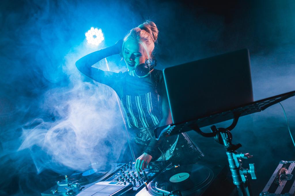 stijlvolle blonde dj meisje aanraken van dj apparatuur en glimlachend in nachtclub met rook  - Foto, afbeelding