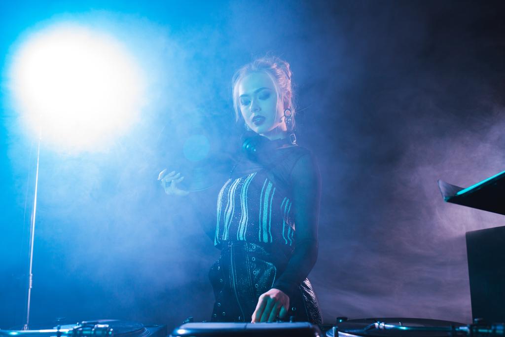 blond dj žena držící retro vinyl záznam a při pohledu na dj vybavení v nočním klubu s kouřem  - Fotografie, Obrázek