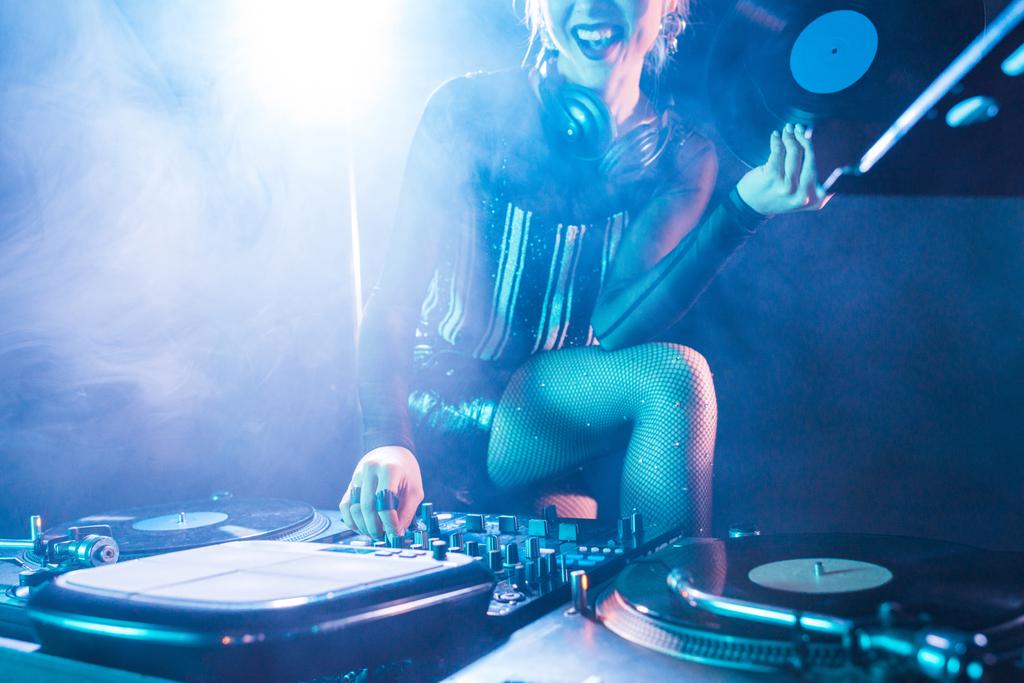 vista recortada de la alegre mujer dj celebración de disco de vinilo retro y de pie cerca de equipo dj en discoteca con humo
  - Foto, Imagen