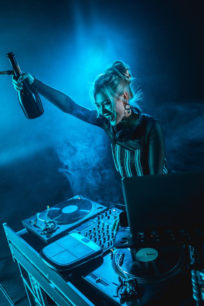 şişe tutan ve duman ile gece kulübünde dj ekipmanları yakınındaki gülümseyen neşeli sarışın dj kadın - Fotoğraf, Görsel
