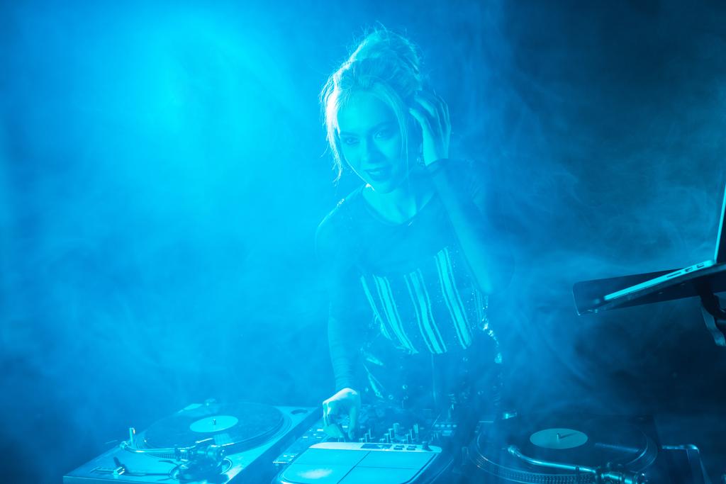 fröhliches blondes DJ-Mädchen hört Musik in Kopfhörern in der Nähe von DJ-Geräten in einem Nachtclub mit Rauch  - Foto, Bild