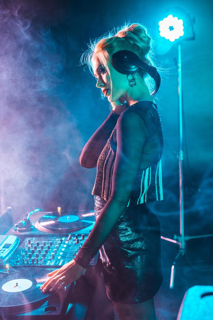 beautiful dj woman with blonde hair listening music and holding headphones in nightclub with smoke - Photo, Image