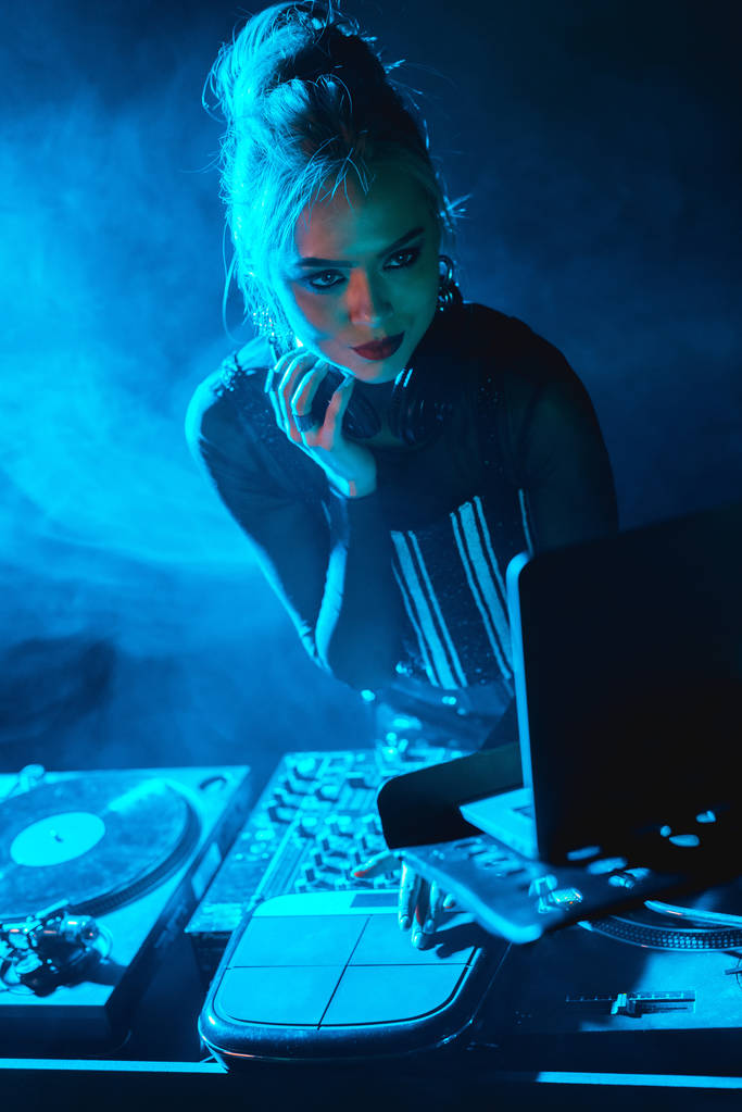 серьезная диджейка с светлыми волосами смотрит на ноутбук в ночном клубе с дымом
   - Фото, изображение